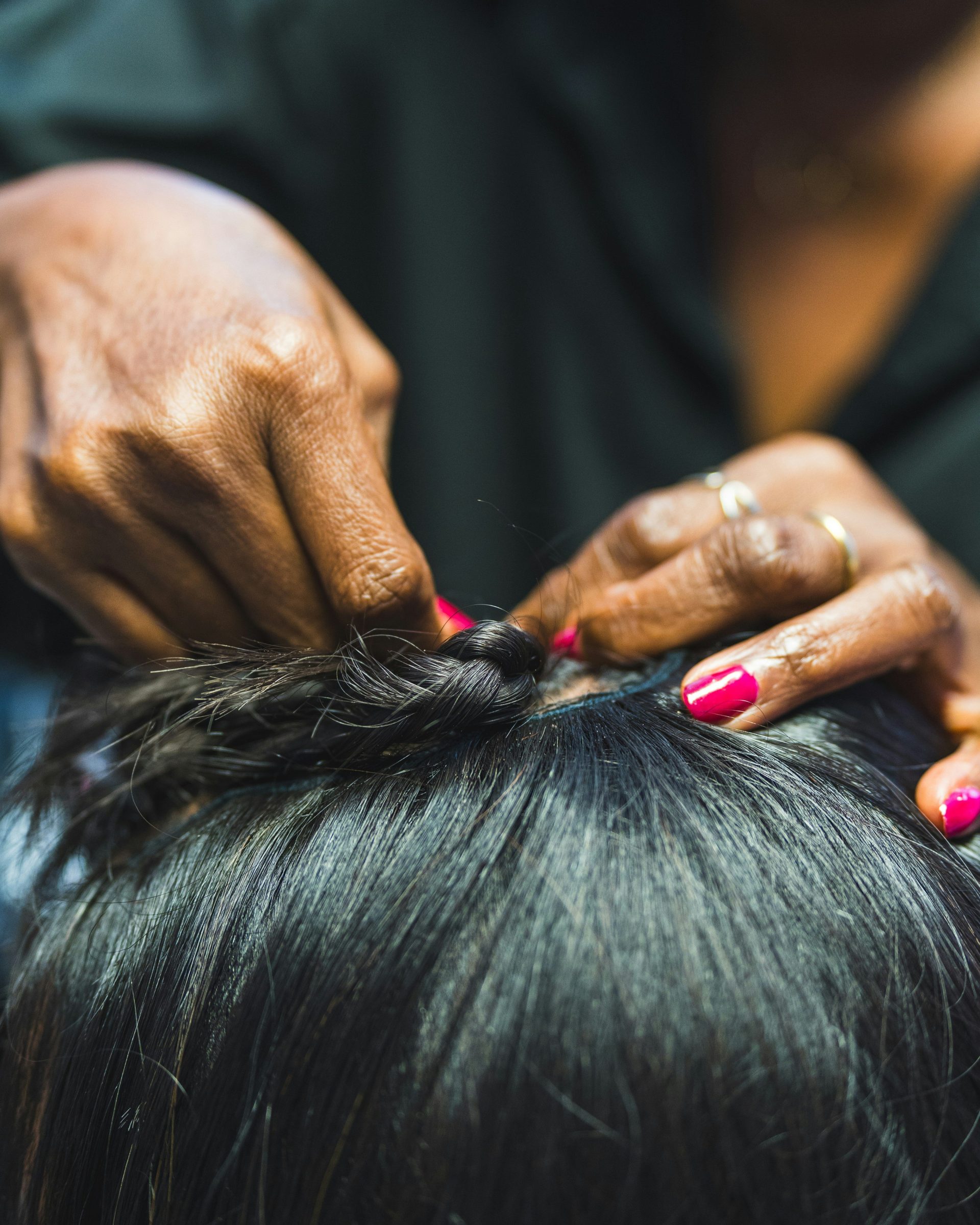 Des mains tressent des cheveux foncés avec des ongles roses.